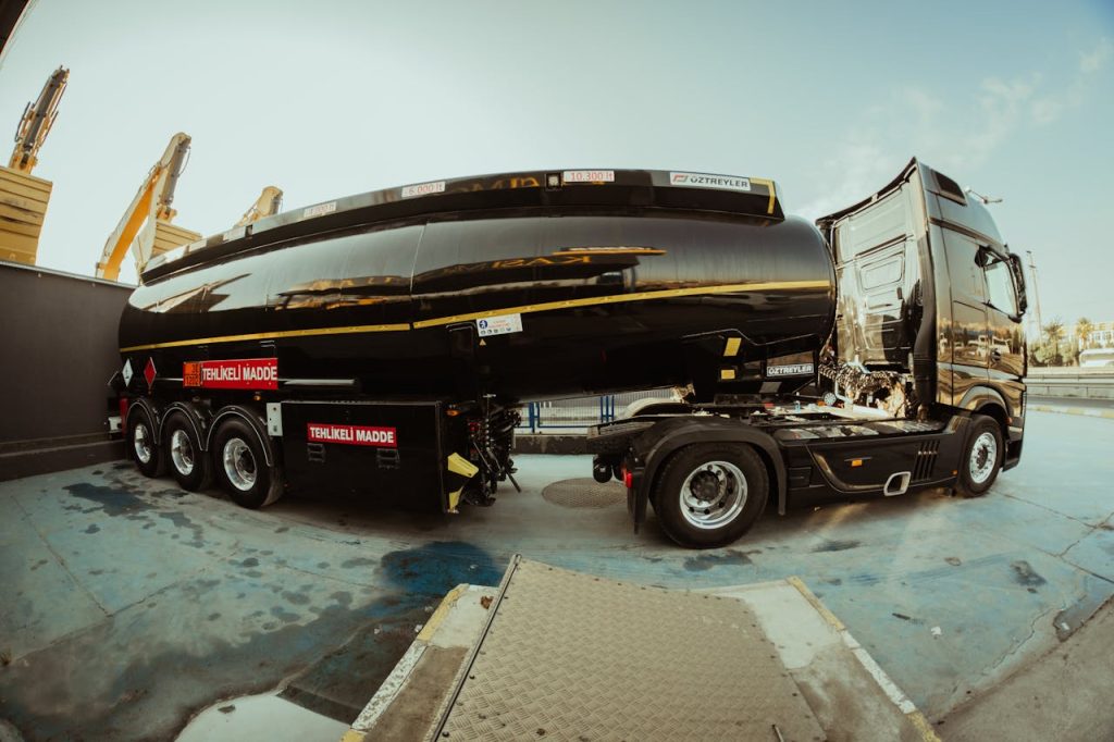 Side view of a black tanker truck parked at an industrial shipping facility under clear skies.